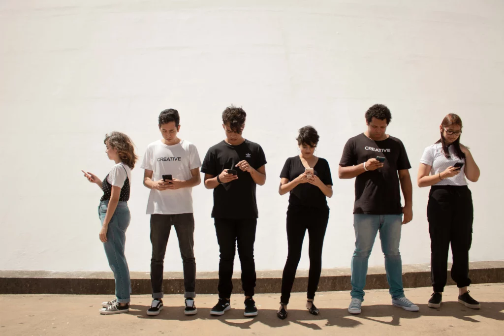 Six young adults stand next to each other, all looking at their phones.