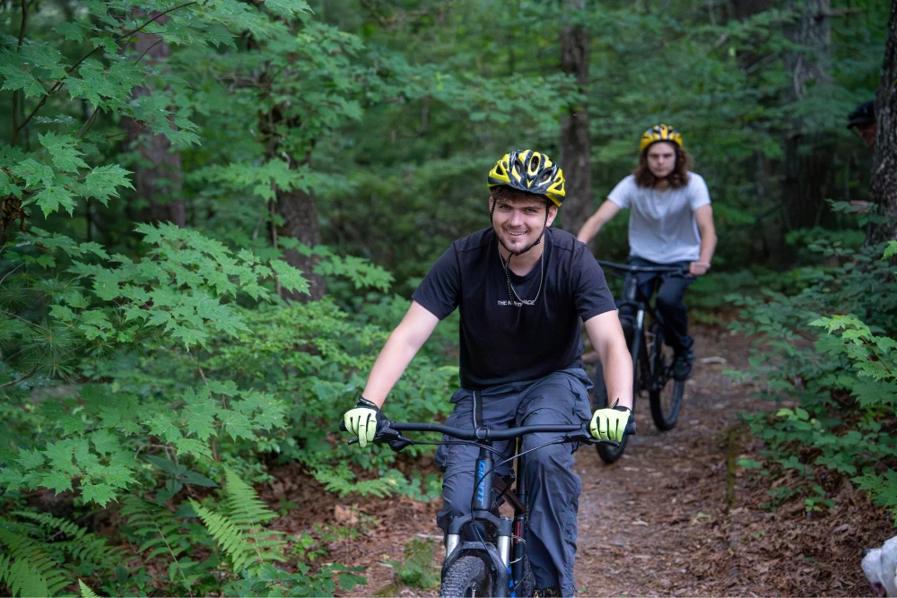 Two young adults ride on a narrow trail through a lush wooded area. The lead cyclist is smiling, wearing a black t-shirt. Both wear yellow helmets