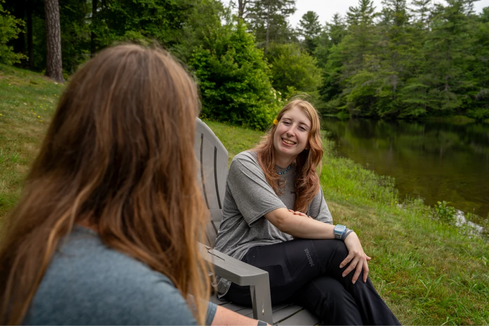 A student with a flower in her hair is smiling and conversing with another person by the lake.