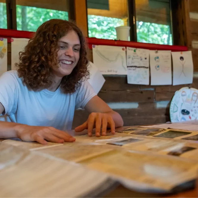 A young adult with curly hair smiles as he looks at a large map on the table. The background shows drawings and crafts displayed on the wall of a wooden cabin.