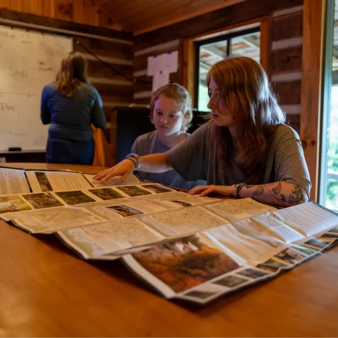 Two young women examine a large map on a table inside a wooden cabin with a lot of natural lighting. In the background, a teacher stands in front of a whiteboard.