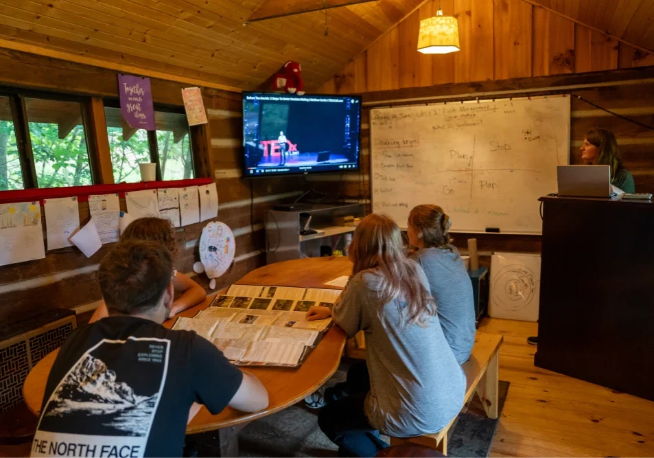A group of young adult students watches a TEDx video on a big modern screen in a rustic classroom with wooden walls. Notes and diagrams about decision-making are written on a whiteboard.