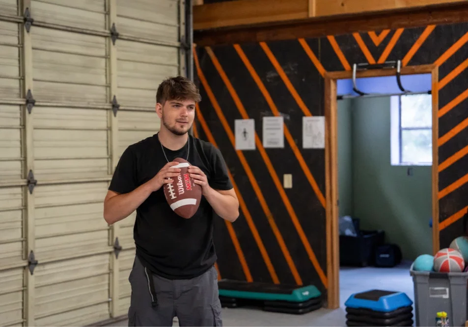 A young man holds a football in a gym with orange and black walls. Exercise equipment is visible in the background.