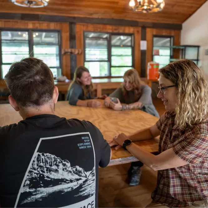 Two pairs of young adult students and their therapists sitting around a wooden table inside a cabin, engaged in conversation about technology. One student plays the guitar.